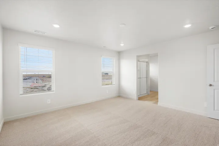 Recessed lighting, light-colored carpeting, multiple windows with blinds, a white paneled door, and an entryway to a bathroom with a glass shower enclosure