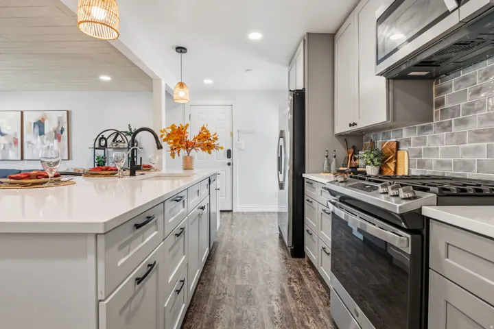 Kitchen featuring a large island with an integrated sink, light-toned countertops, gray cabinetry, stainless steel appliances, and a gray subway tile backsplash