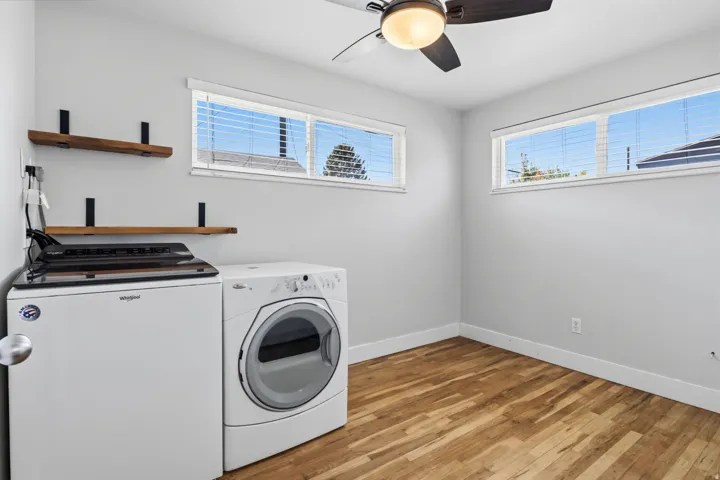 Light gray walls, wood-finish flooring, two large windows with blinds, a ceiling fan, and two wall-mounted wood shelves