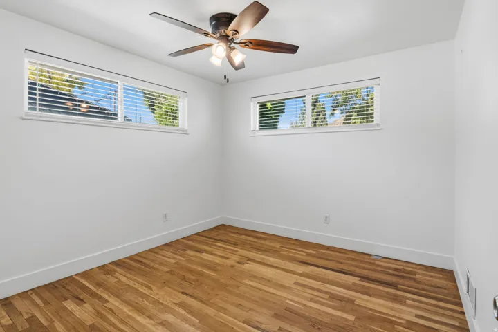 Hardwood flooring, white walls, two horizontal windows with blinds, and a ceiling fan with integrated lighting