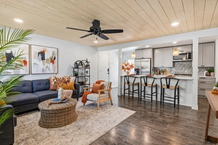 Wood-finish flooring throughout, light wood-paneled ceiling with recessed lighting and ceiling fan, and a kitchen featuring light gray cabinetry and stainless steel appliances