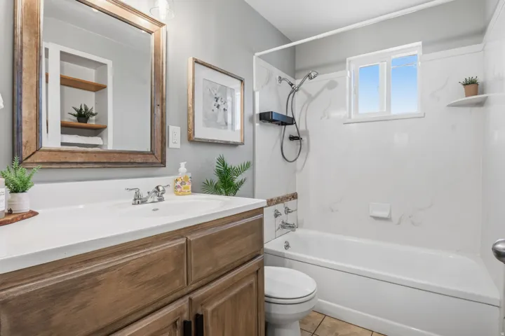 Bathroom featuring a wood-finish vanity with an integrated sink, a large framed mirror, a tub-shower combination with white surround, and a compact window