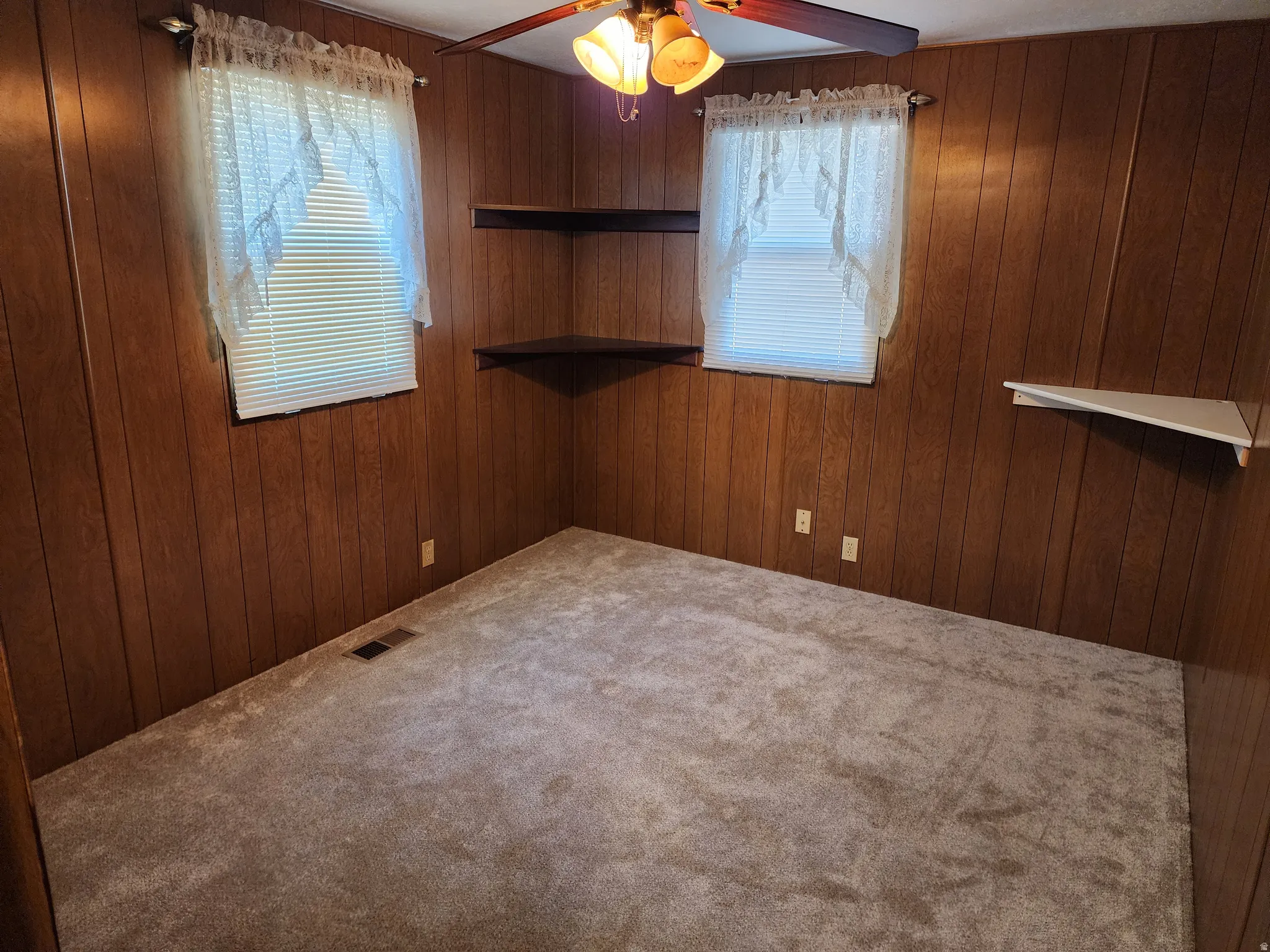 Wood-paneled walls, two windows with blinds, built-in corner shelving, overhead ceiling fan with light fixture, and carpeted flooring
