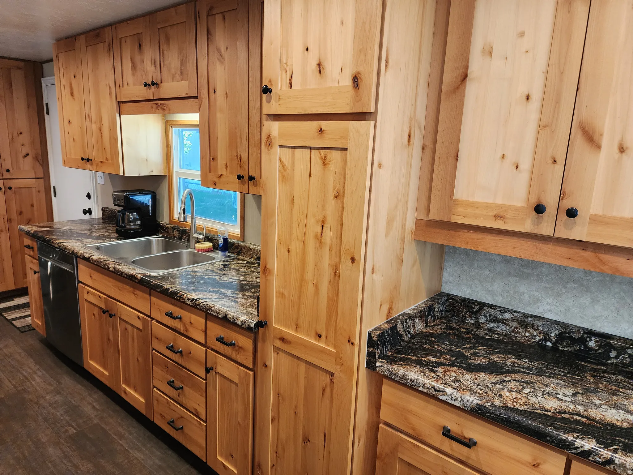 Natural wood cabinetry, dark patterned countertops, stainless steel double basin sink, and black appliances