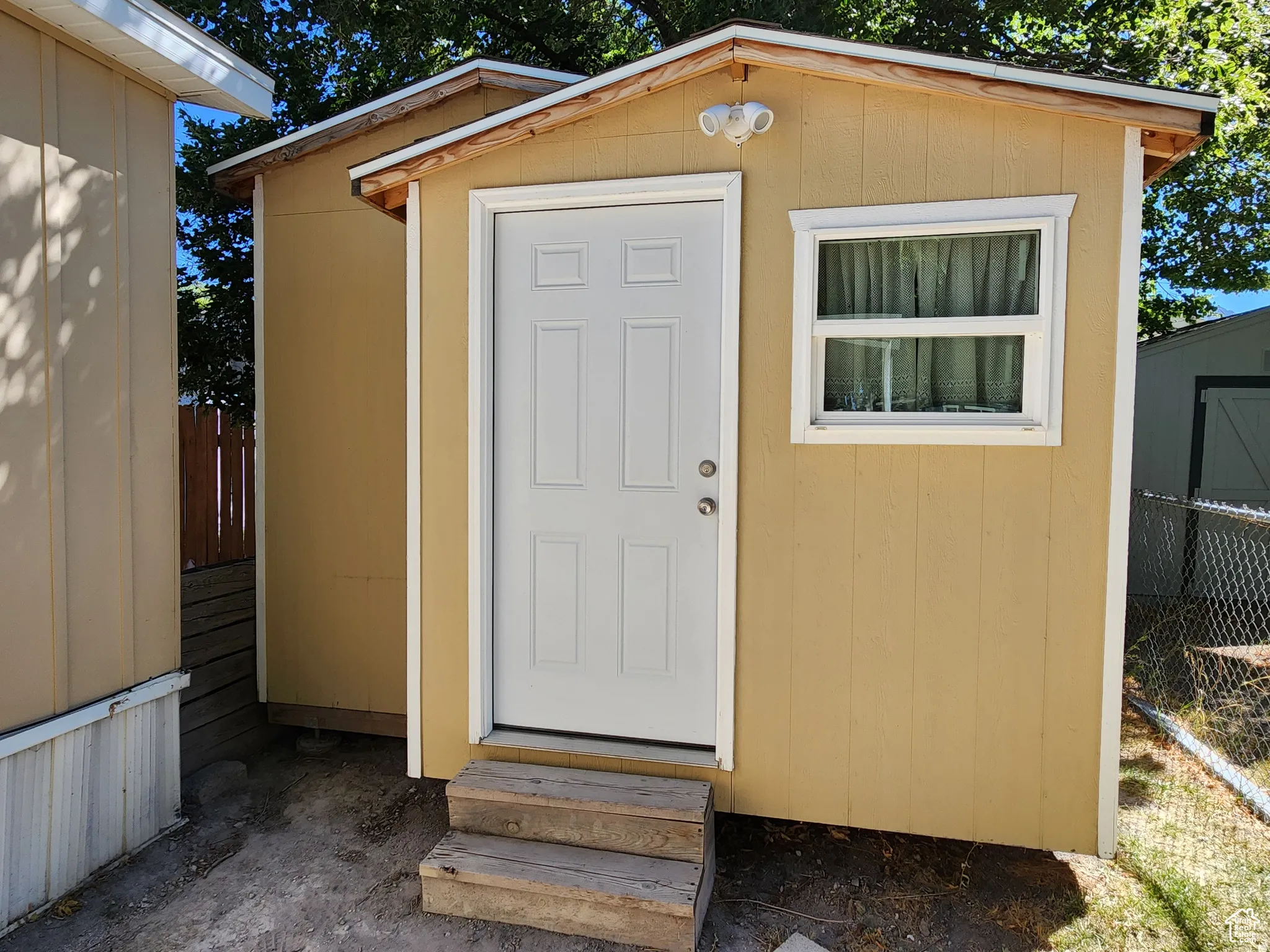 Yellow-painted shed with a white paneled door, a white-framed window, and exterior illumination