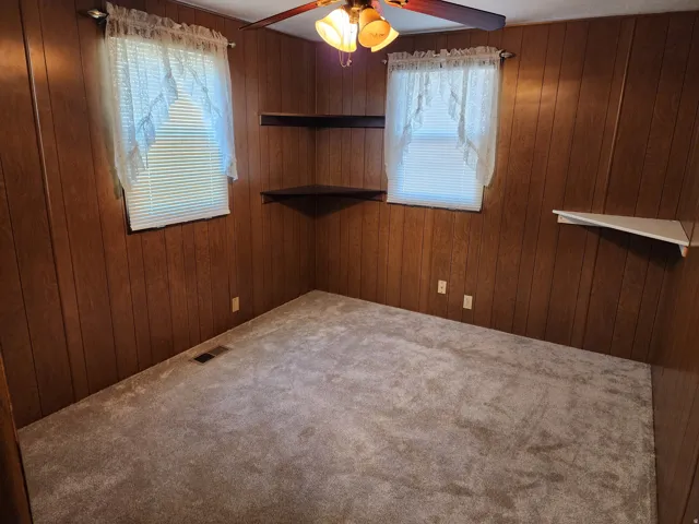 Wood-paneled walls, two windows with blinds, built-in corner shelving, overhead ceiling fan with light fixture, and carpeted flooring