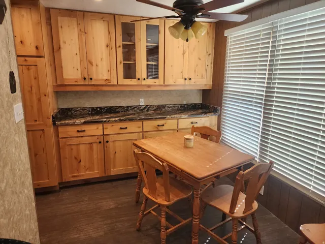 Wood-finish cabinetry with glass-front accents, dark countertop, wood-finish flooring, and a ceiling fan with integrated lighting