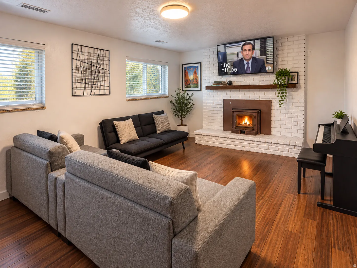 Wood-finish flooring throughout, white brick fireplace with wood mantel, recessed wood-burning insert, and multiple windows with horizontal blinds