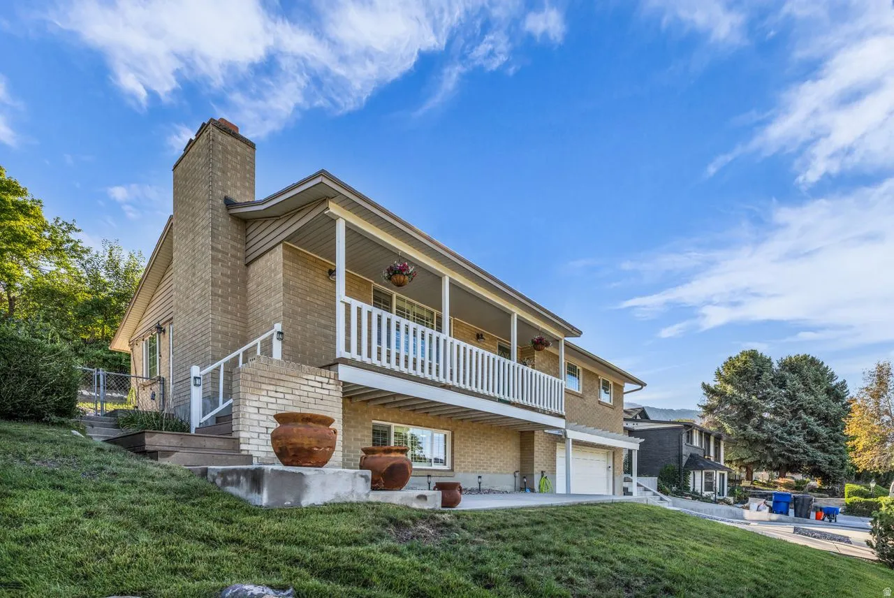 Brick exterior with a prominent chimney, upper-level balcony with white railings, lower-level patio, and an integrated garage