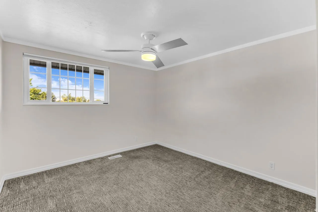 Neutral wall paint, ceiling fan with integrated lighting, window with grid inserts, and textured carpet flooring