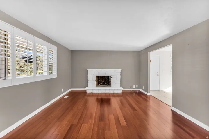 Wood-finish flooring, white stone fireplace, multiple windows with plantation shutters, and light-toned walls