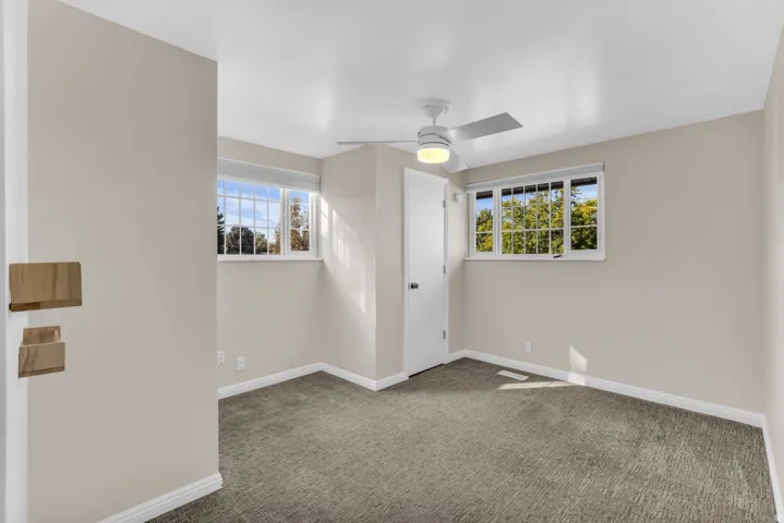 Neutral wall paint, carpeted flooring, two windows with grilles, and a ceiling fan with light fixture