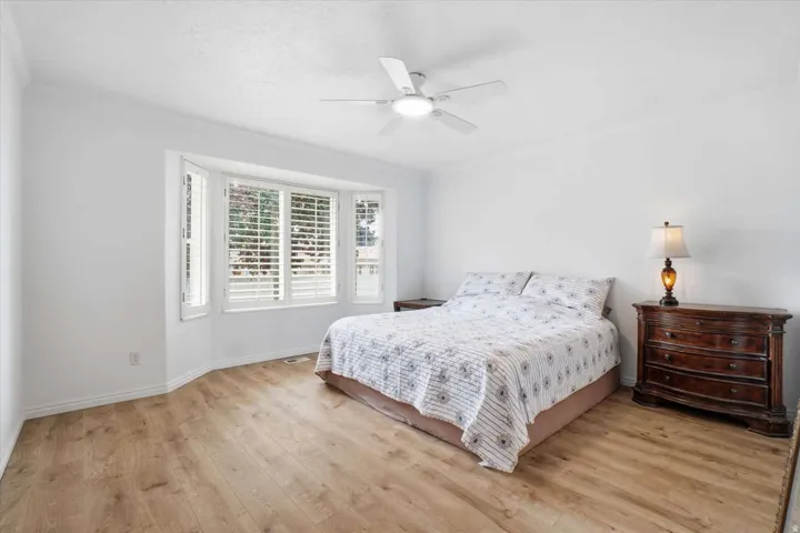 Bay window with plantation shutters, wood-finish flooring, ceiling fan with integrated lighting, and crown molding