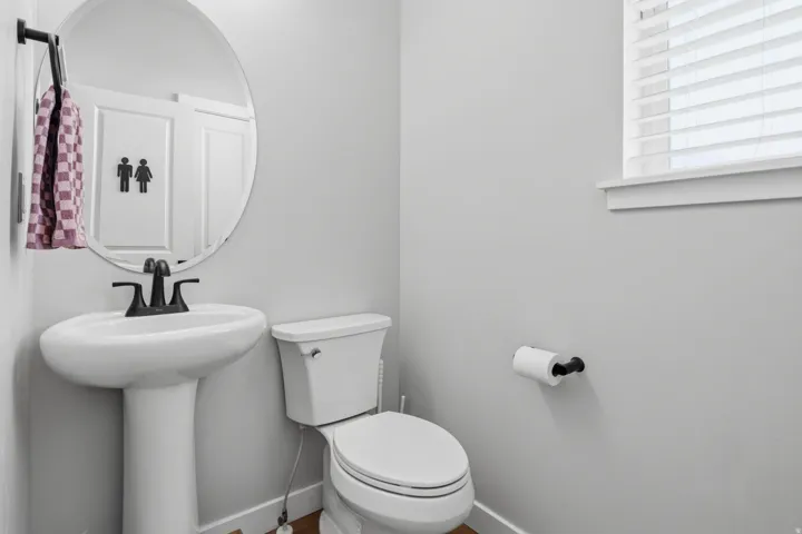 Pedestal sink with a matte black faucet, round wall-mounted mirror, and a window with blinds