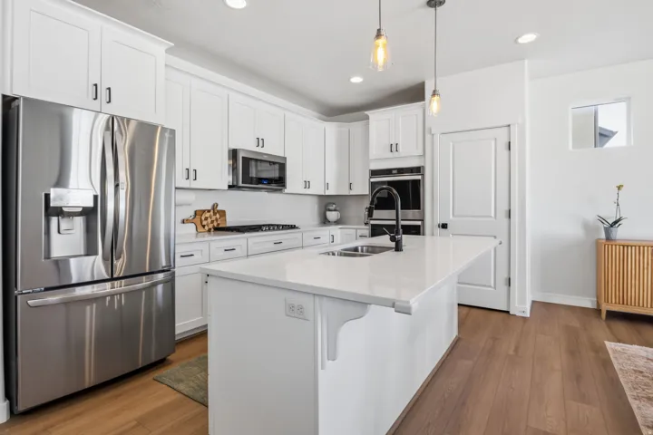 White shaker cabinetry, stainless steel appliances, central island with integrated sink, wood-finish flooring, and pendant lighting