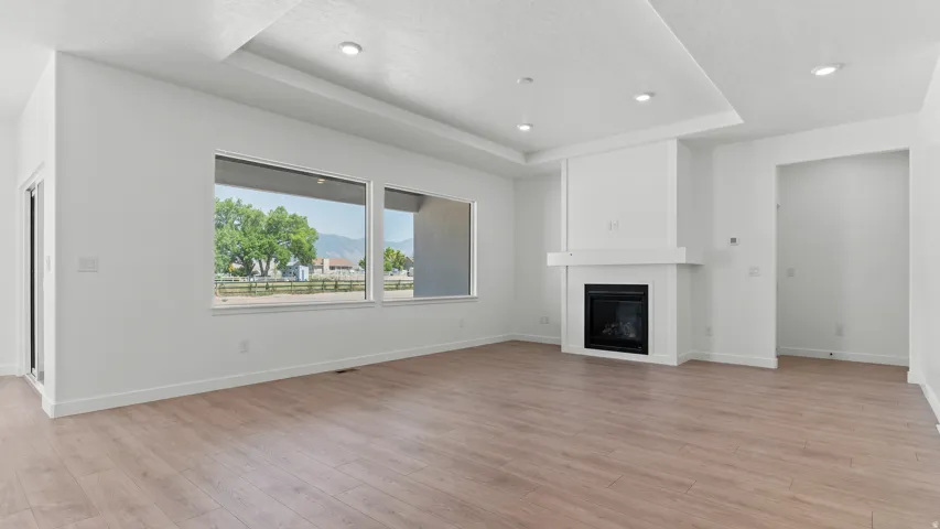 Spacious room featuring wood-finish flooring, a contemporary fireplace with a white mantel, a tray ceiling with recessed lighting, and large windows