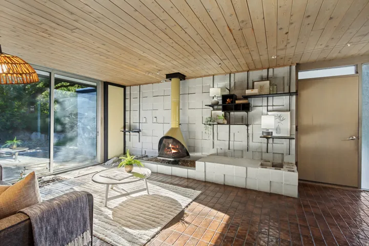 Tongue and groove wood panel ceiling, floor-to-ceiling sliding glass doors, suspended fireplace with stone hearth, geometric wall paneling, and ceramic tile flooring