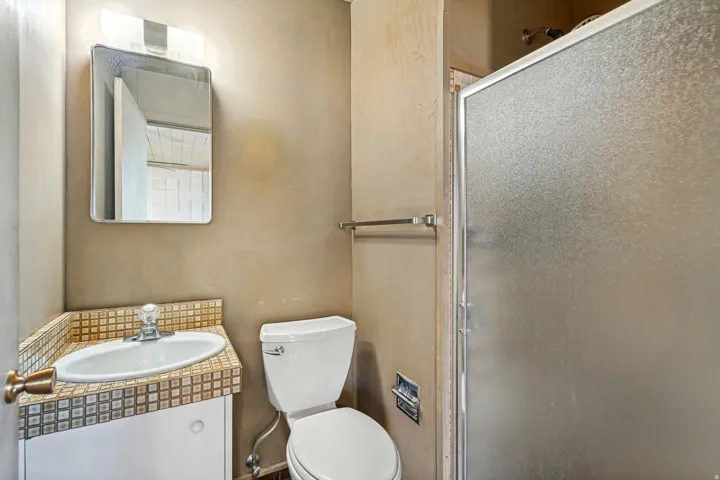 Vanity with integrated sink, mosaic tile countertop, wall-mounted mirror, and glass-enclosed shower