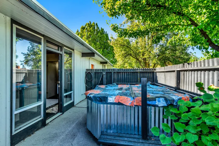 Sliding glass door entry, concrete patio, corrugated metal fencing, mature trees, and landscaping