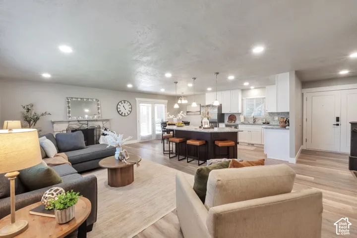 Open-concept living space featuring wood-finish flooring, a stone-faced fireplace, a kitchen island with pendant lighting, white cabinetry, and double French doors