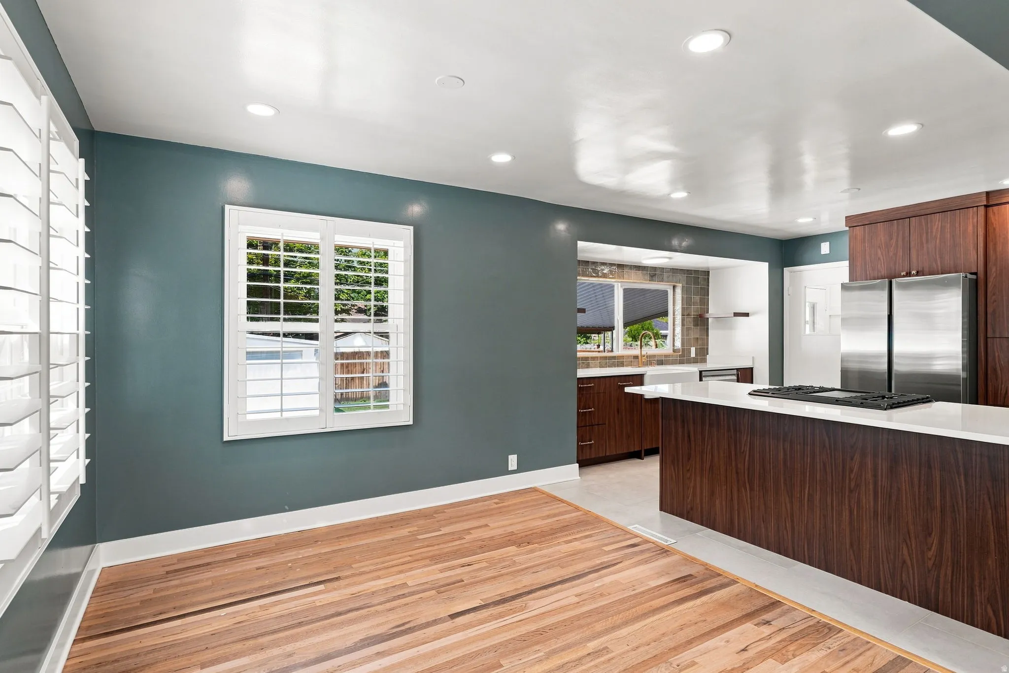 Hardwood flooring, white window shutters, recessed ceiling lighting, kitchen island with integrated cooktop, and stainless steel refrigerator