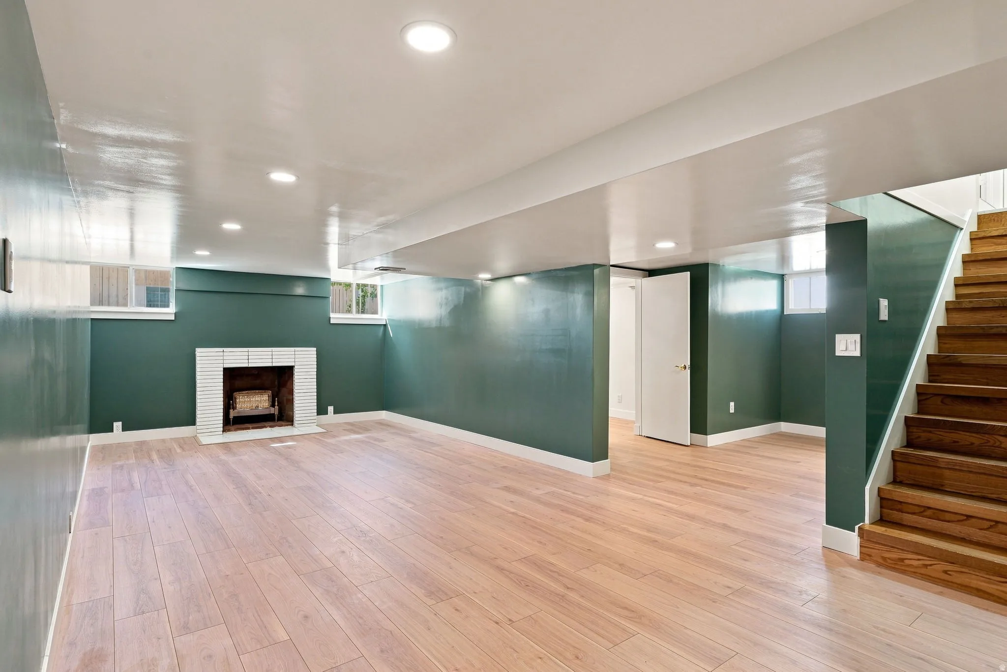 Spacious room featuring wood-finish flooring, a white brick fireplace, recessed lighting, and a wood staircase