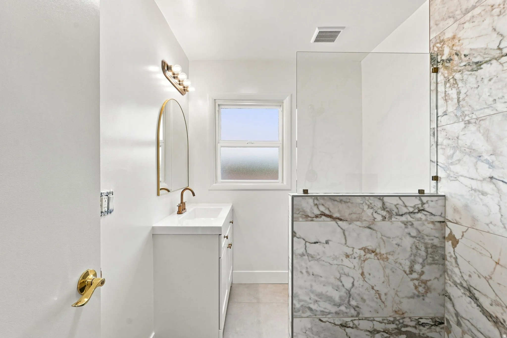 White vanity with integrated sink, brass fixtures, arched mirror, and a walk-in shower with marble-look wall tiling and a glass enclosure