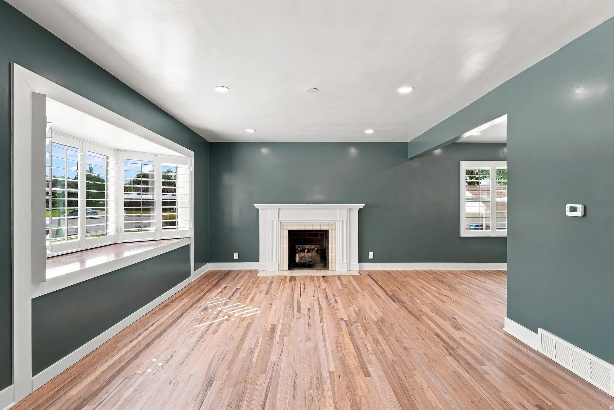 Hardwood flooring, bay window with plantation shutters, recessed lighting, and a fireplace with a detailed mantel