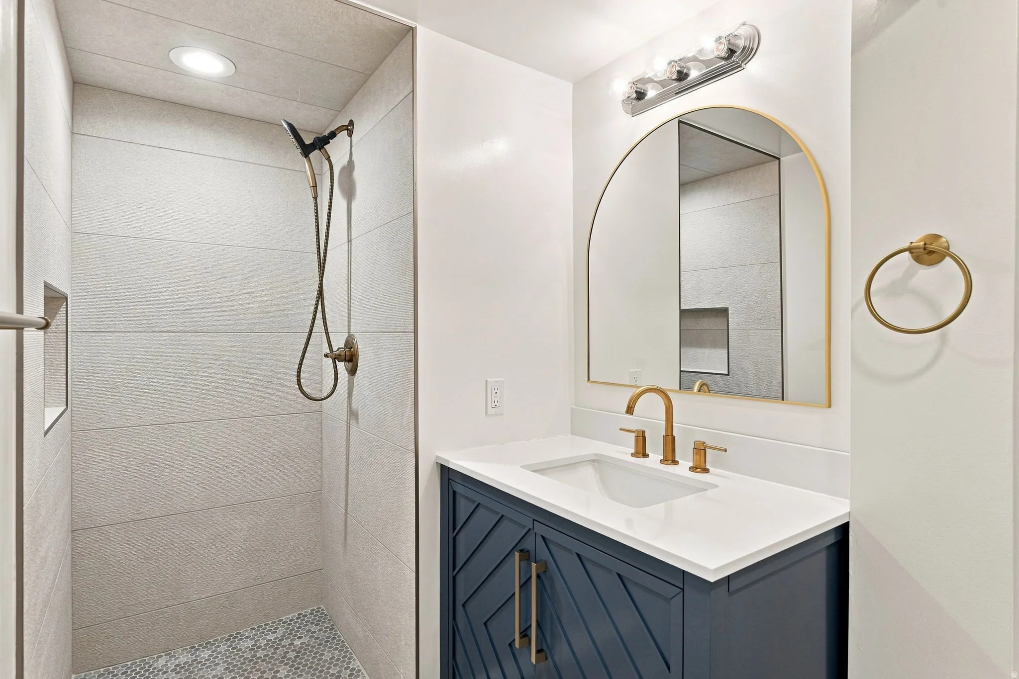 Contemporary bathroom featuring a navy blue vanity with gold hardware, a white countertop, an arched gold-framed mirror, and a tiled walk-in shower with a recessed niche
