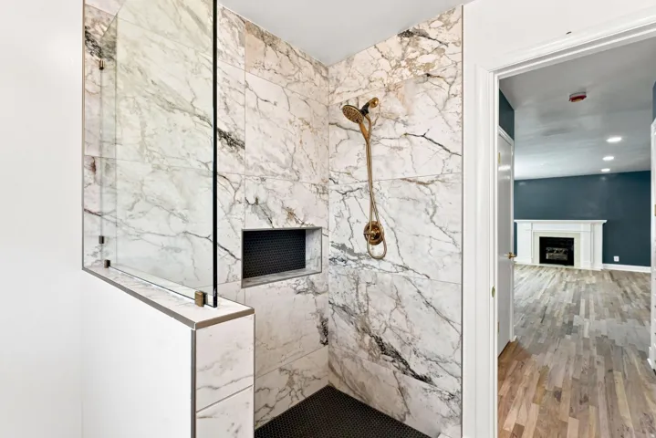 Walk-in shower with full-height marble-patterned tile, glass partition, recessed shower niche, and a bronze-finish shower system