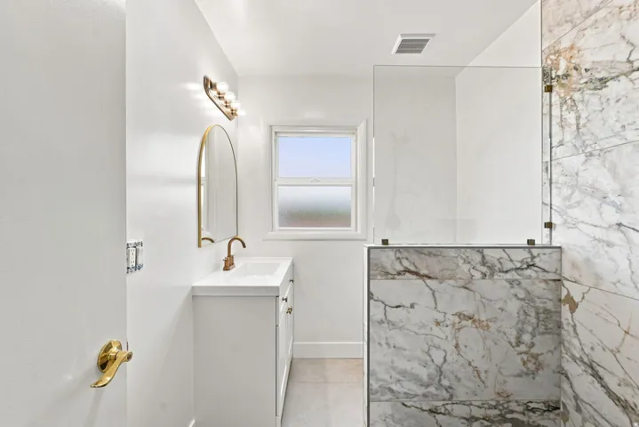 White vanity with integrated sink, brass fixtures, arched mirror, and a walk-in shower with marble-look wall tiling and a glass enclosure