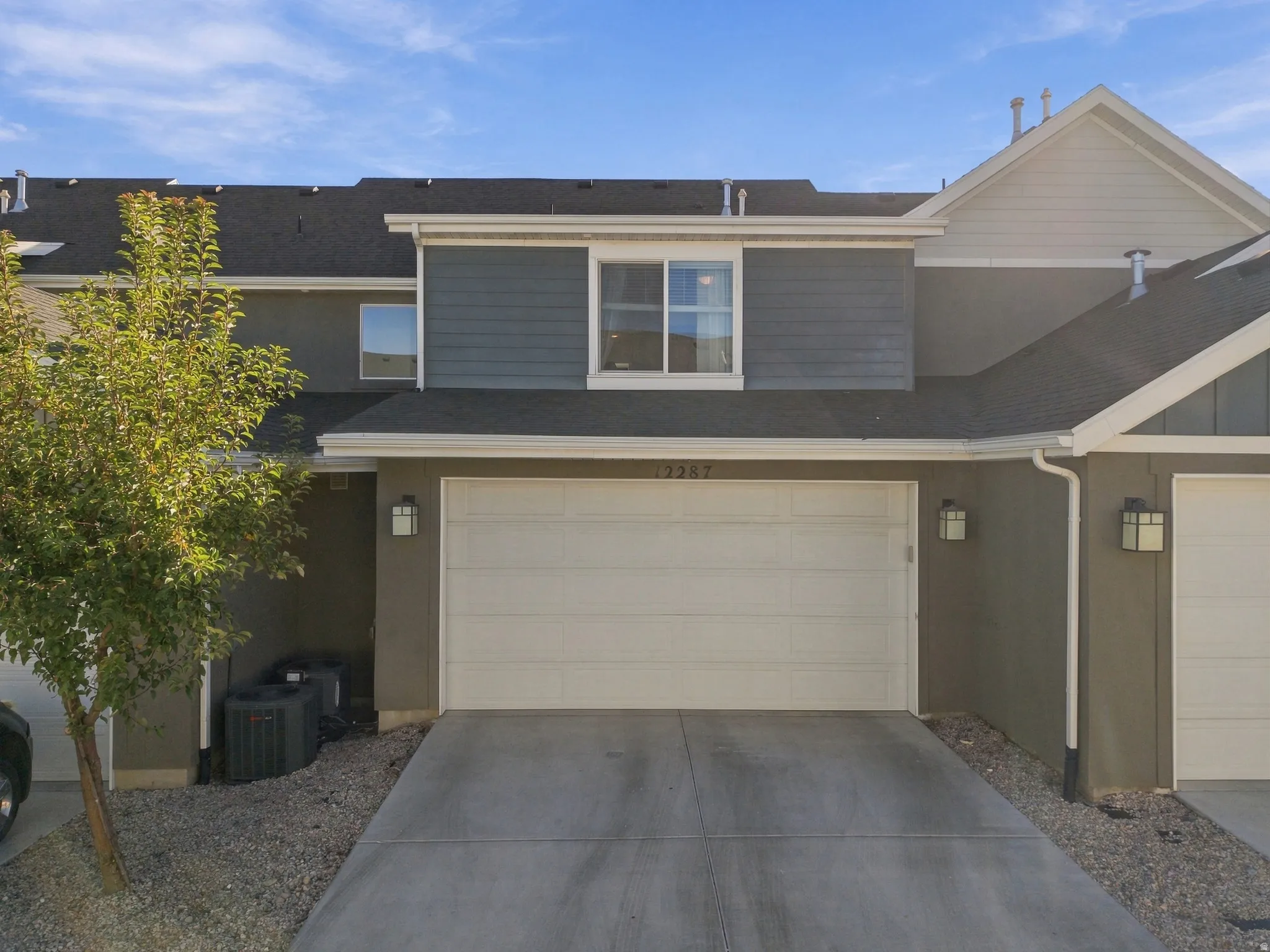 Attached two-car garage, concrete driveway, dark gray exterior siding, light gray shingle roof, and exterior lighting fixtures