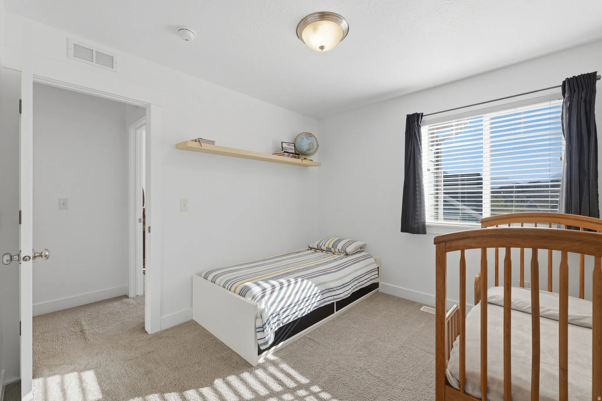 White walls and ceiling, wall-mounted shelving, neutral carpet flooring, single window with blinds, flush mount ceiling light fixture