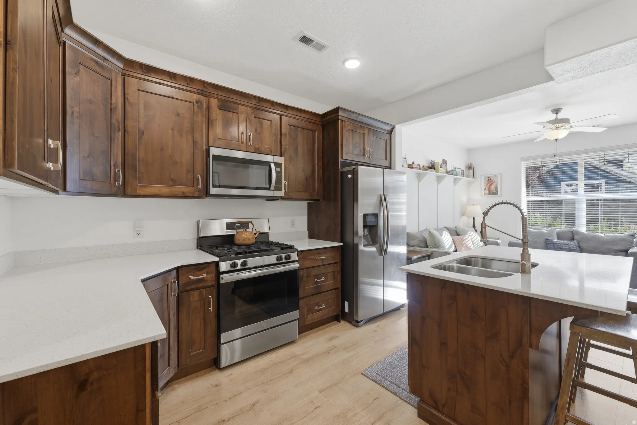 Kitchen featuring wood cabinetry, stainless steel appliances, light-toned countertops, a kitchen island with a double basin sink, and wood-finish flooring