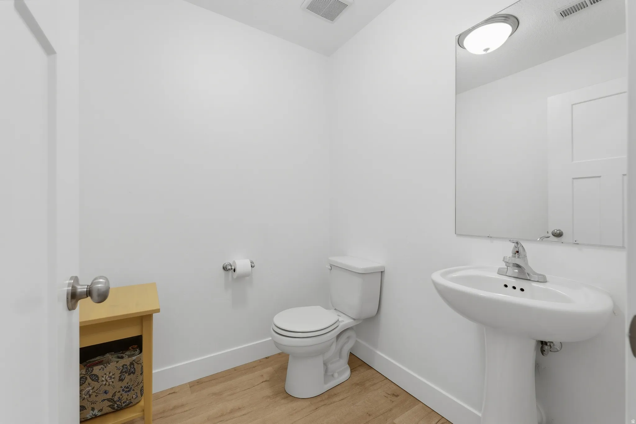 White pedestal sink with a chrome faucet, wall-mounted mirror, and wood-finish flooring