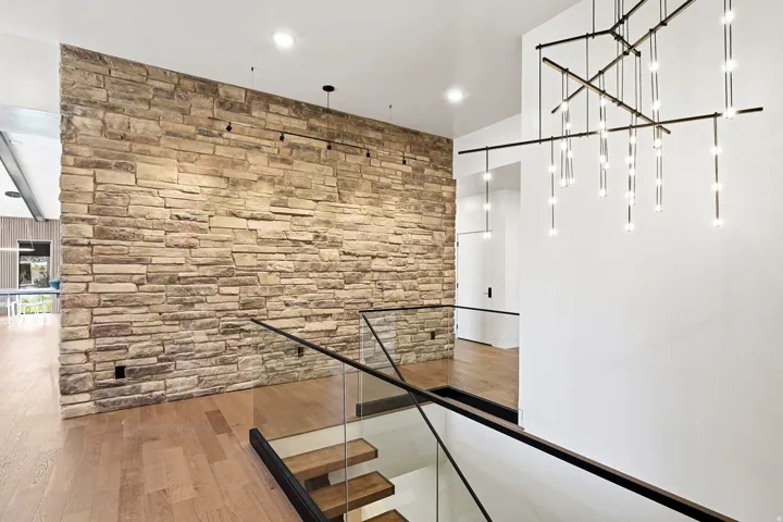 Stone-clad accent wall, glass panel railing with dark wood-finish top rail, wood-finish flooring, and a multi-light linear chandelier
