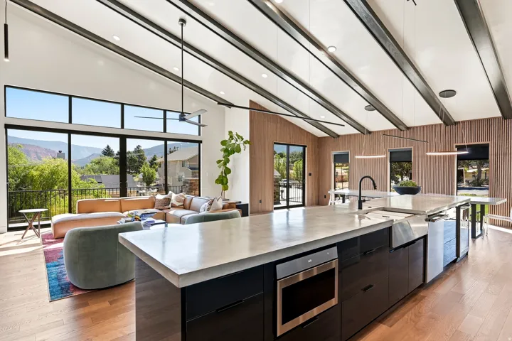 Vaulted ceiling with exposed beams, wood-finish flooring, full height windows, large kitchen island with integrated appliances, and a modern ceiling fan