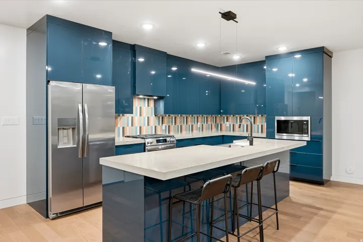 Contemporary kitchen with high-gloss blue cabinetry, stainless steel appliances, light-toned countertops, and a colorful tile backsplash