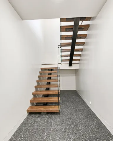 Floating wood-finish staircase with a metal support beam, glass railing, and dark speckled tile flooring
