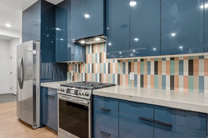 Blue high-gloss cabinetry, multi-colored vertical tile backsplash, stainless steel gas range, light-toned countertop, and wood-finish flooring