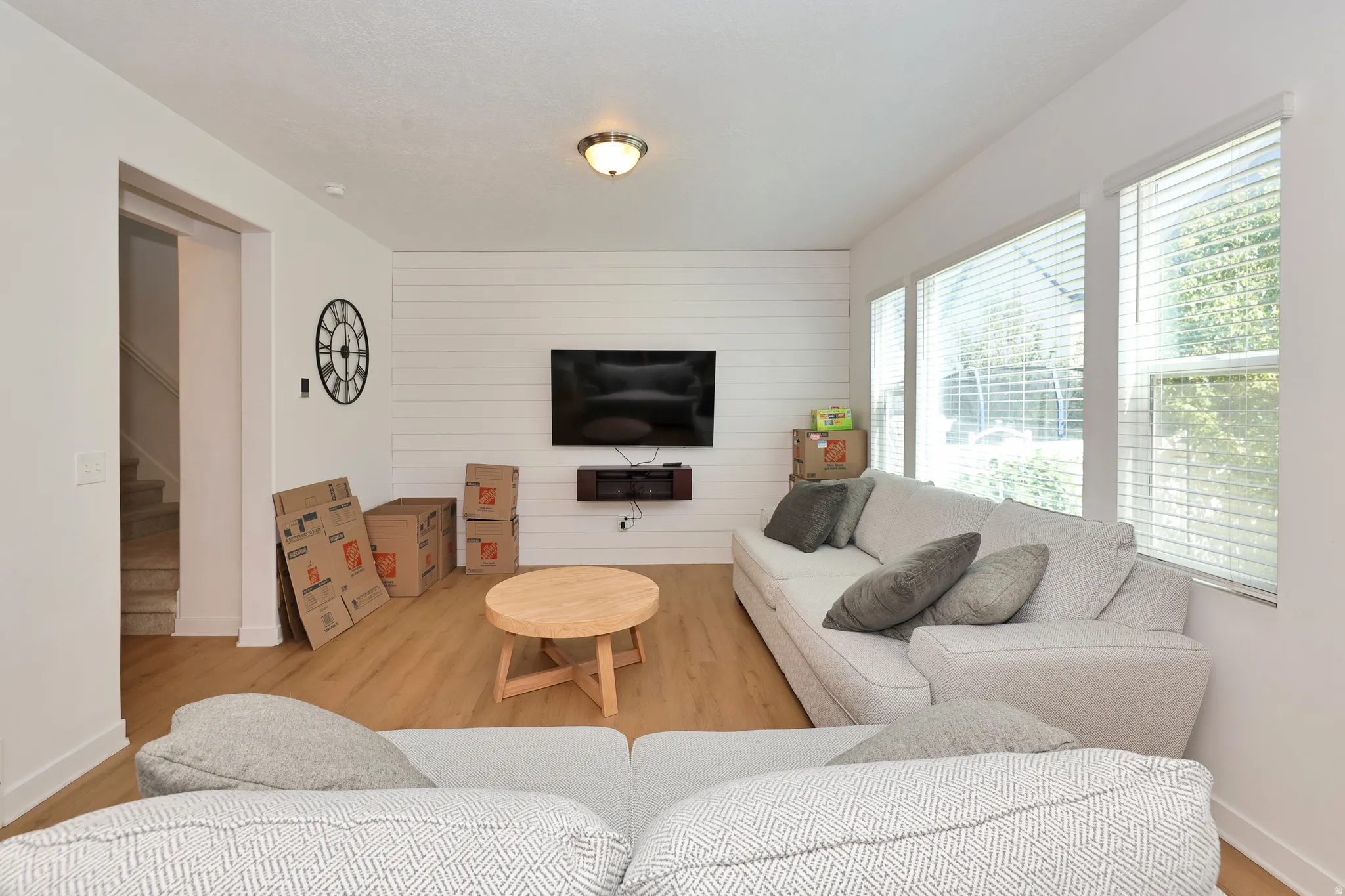 Wood-finish flooring, accent wall with horizontal paneling, multiple large windows with blinds, recessed lighting, and a visible staircase