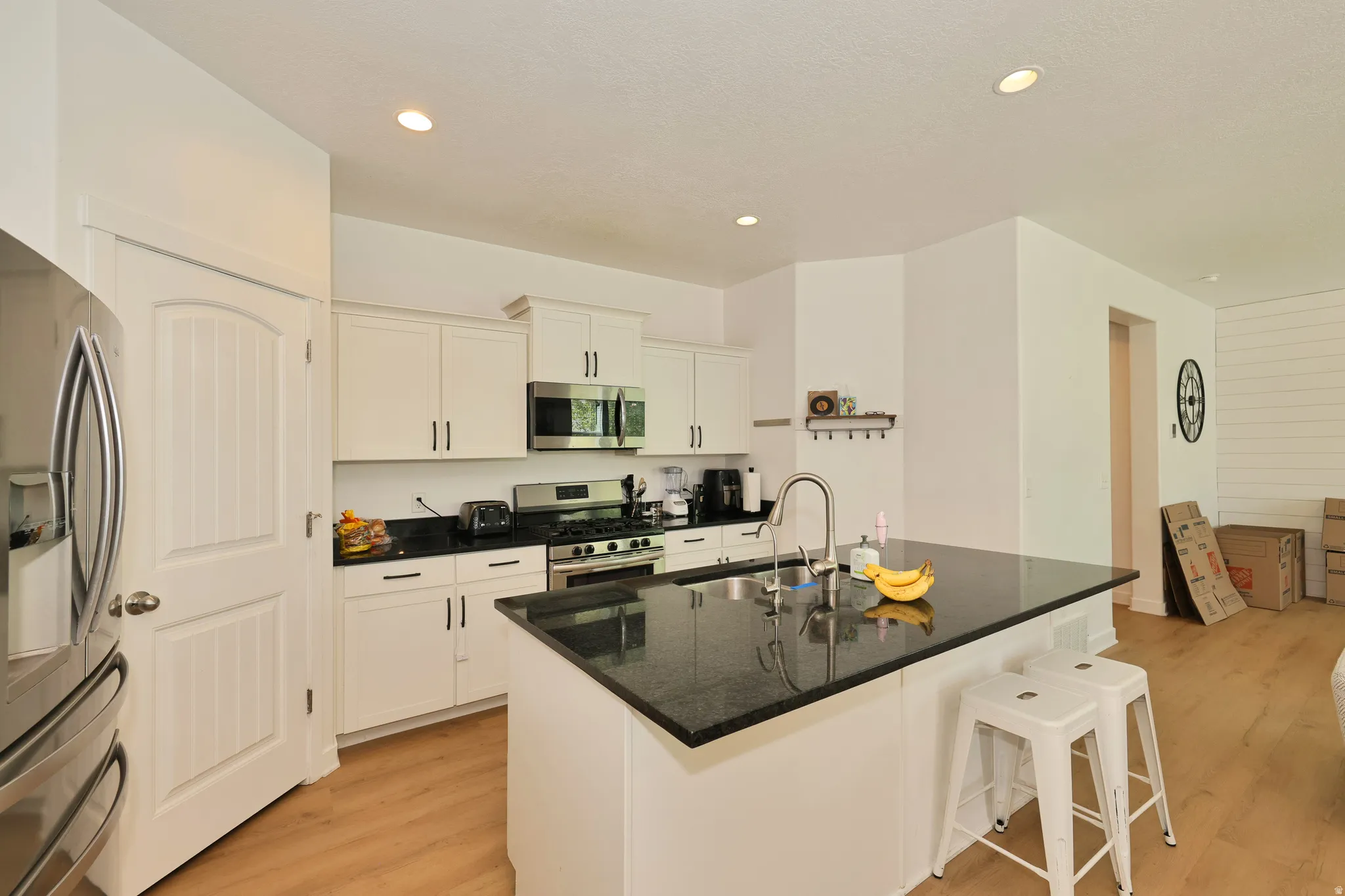 Kitchen with white cabinetry, stainless steel appliances, black countertops, a center island with sink, and wood-finish flooring
