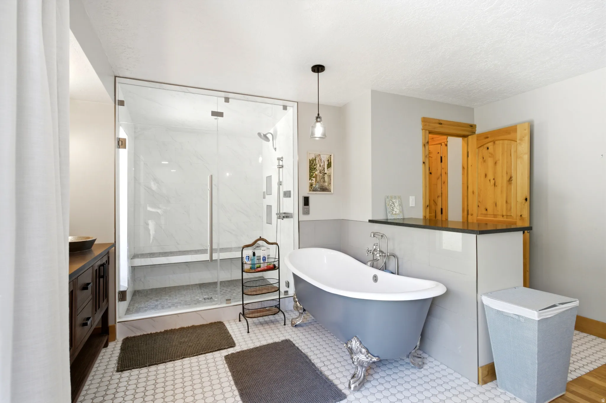 Glass-enclosed shower with a built-in bench, freestanding clawfoot tub, white hexagonal floor tiling, and a dark wood-finish vanity with a vessel sink