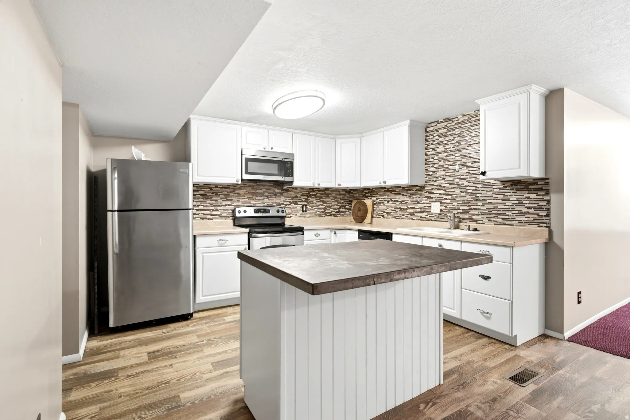 White cabinetry with dark hardware, a mosaic tile backsplash, stainless steel appliances, wood-finish flooring, and a kitchen island with a contrasting countertop