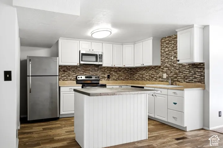 White cabinetry with brushed nickel hardware, stainless steel appliances, patterned tile backsplash, wood-finish flooring, and a kitchen island with a stone-finish countertop
