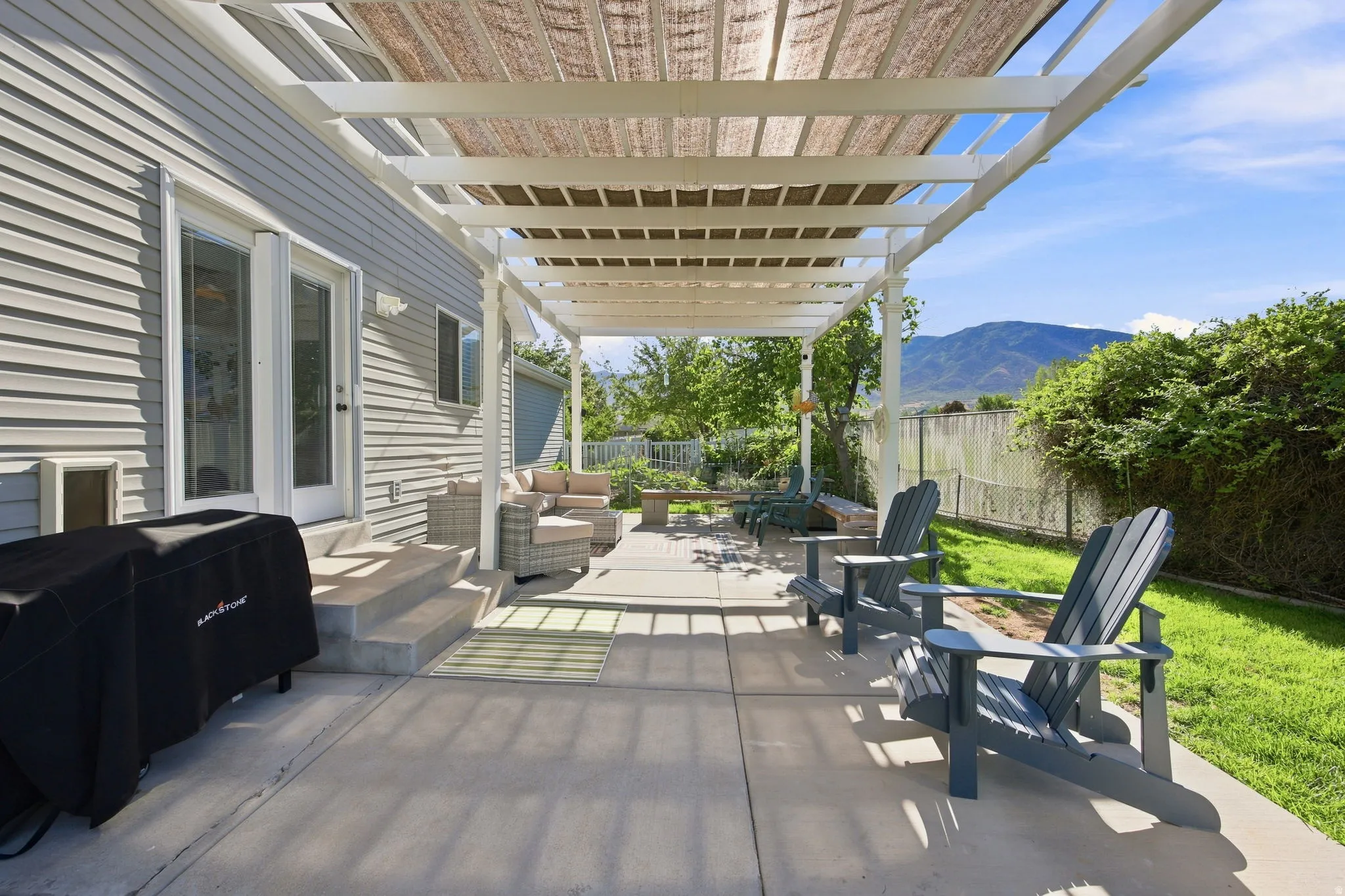 Expansive concrete patio featuring a white pergola with a retractable shade cover, leading to a manicured lawn