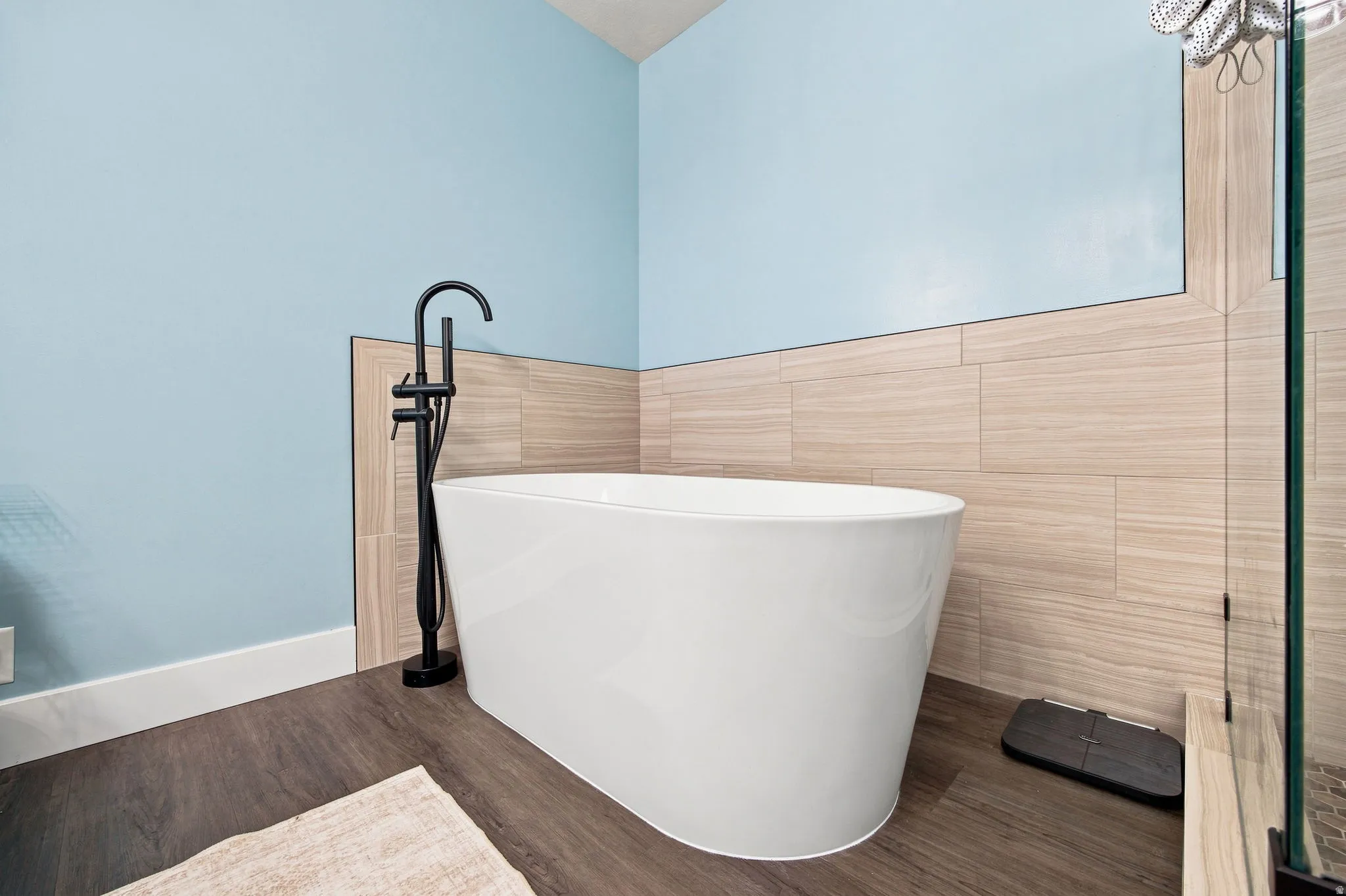 Freestanding soaking tub with a matte black floor-mounted faucet, wood-finish flooring, and light-toned wall tiling
