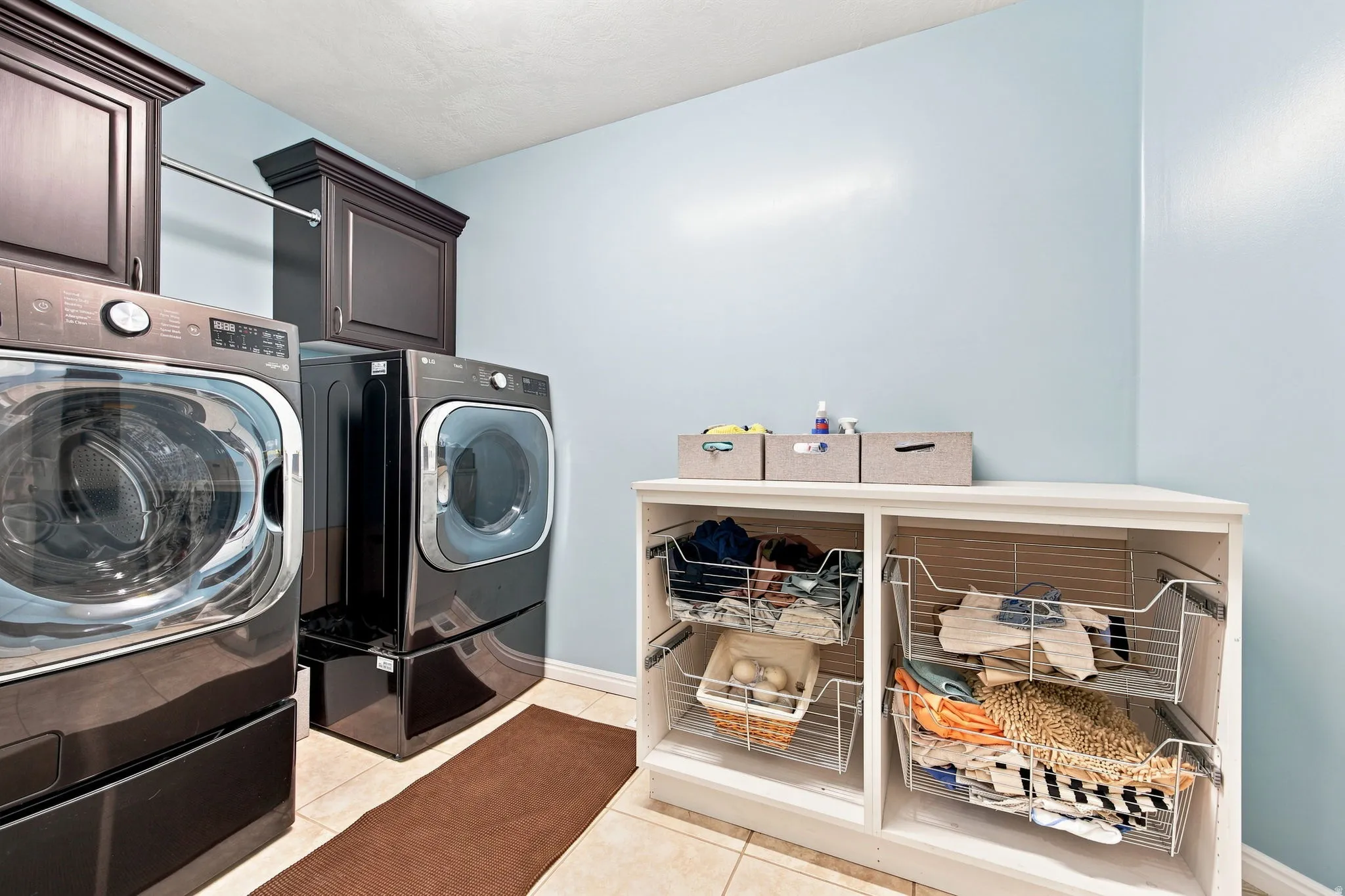 Laundry area featuring overhead cabinetry, a metal hanging rod, tiled flooring, and a built-in storage unit with pull-out wire baskets