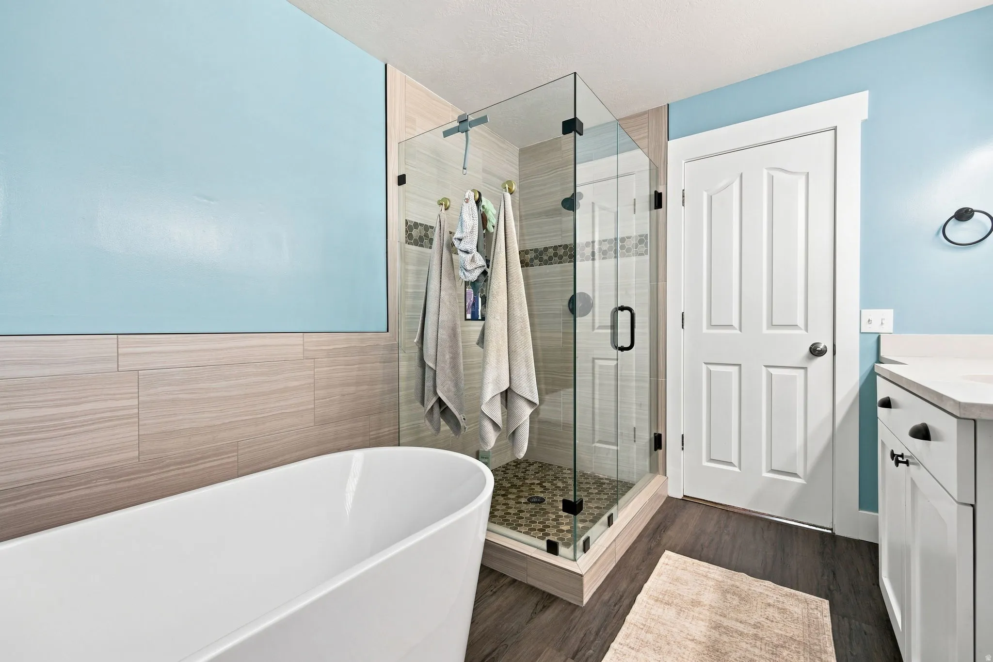 Freestanding soaking tub, glass-enclosed shower with tiled walls and pebble flooring, wood-finish flooring, and a white vanity with a light-colored countertop