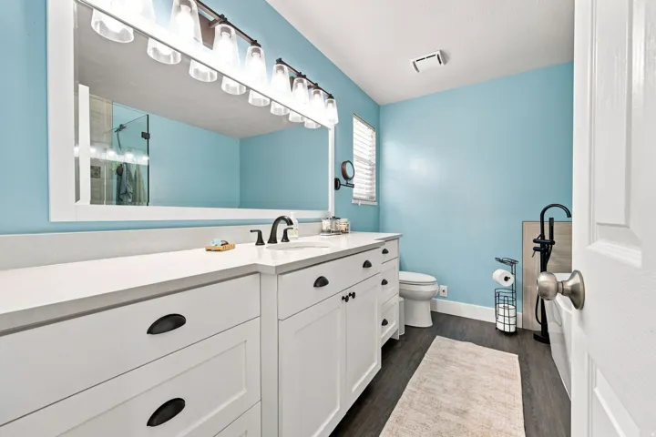Double vanity with white cabinetry and dark hardware, white countertop, large framed mirror, and wood-finish flooring